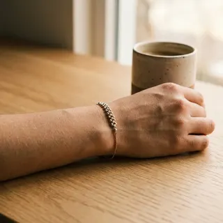 دستبند گوی طلا روی مچ دست کنار فنجان قهوه، با طراحی گوی‌دار و زنجیری ظریف